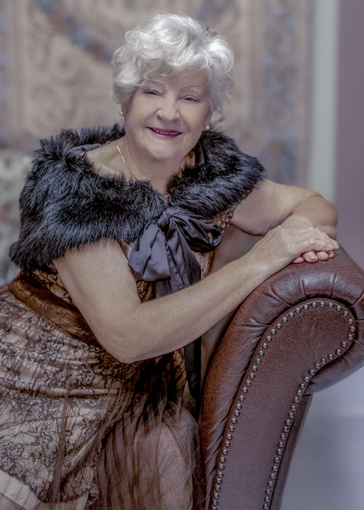 Senior woman in vintage gown and black fur collar sitting on a brown leather couch